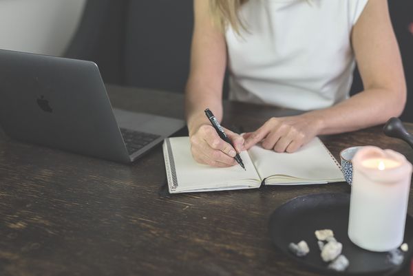 Kvinna i vit tröja skriver i en bok på ett grått skrivbord, där det även står en laptop.